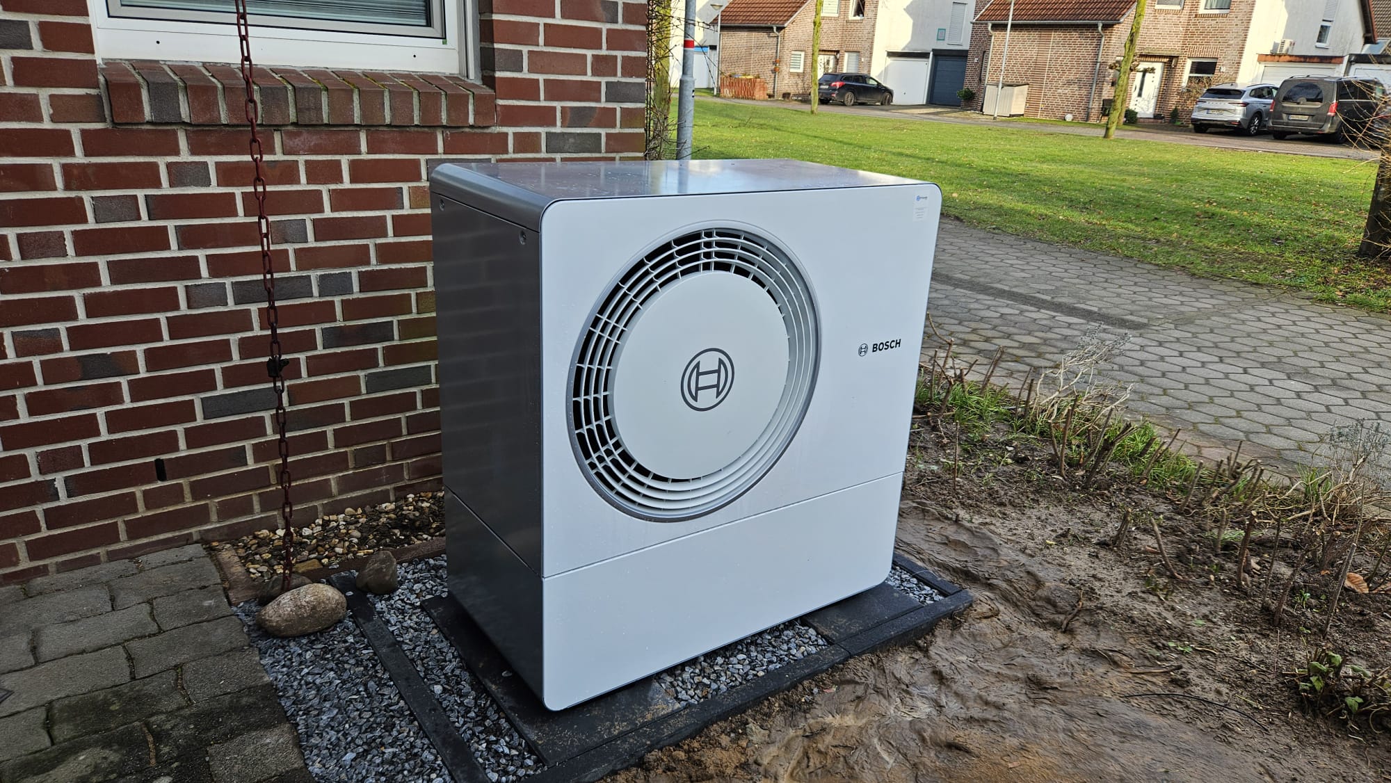 Eine moderne Bosch-Wärmepumpen-Außeneinheit steht sauber installiert auf einem gepflasterten Fundament vor einem Wohnhaus.